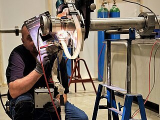 Mann mit Schutzschild in Hockstellung beim Schweißen an einer Hydraulikleitung